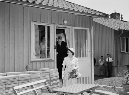 Ett trähus under uppförande. tre peroner står i bakgrunden och tittar på när drottning Elizabeth II och Gustav VI Adolf kliver ut ur huset.