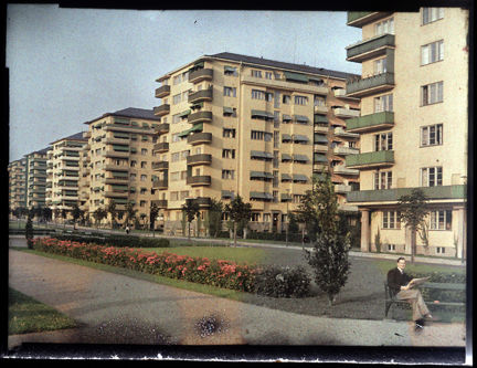 En man läser tidningen på en parkbänk. Funkishus i bakgrunden. Anlagda rabatter med planteringar och träd.