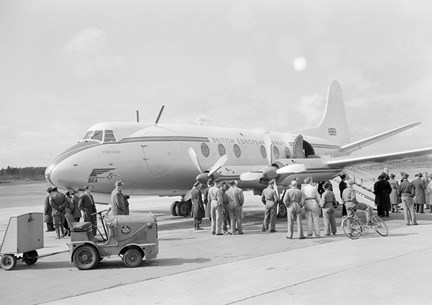 Ett större propellerplan står parkerat på marken framför flygterminalen. Nedanför planet står ett tjugotal människor, en del kabinpersonal, mekaniker och civilklädda. 