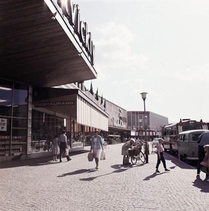 Trottaren utanför Farsta centrum. Människor promenerar och bredvid står en bil och en buss parkerad.