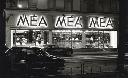 Svartvitt bild på butiksexteriör med neonljus, skyltfönster och bilar. 