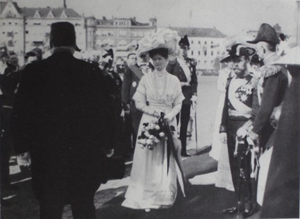 I fokus Alexandra i vit klänning och stor vit hatt. Män i uniform stående runtomkring. Vatten i bakgrunden.