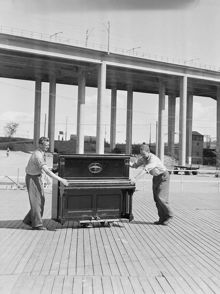 Skanstull. Förberedelser för kommunal parkdans i Eriksdalslunden. Två män rullar ett piano, Skanstullsbron i fonden