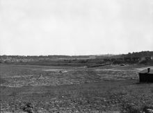 Vy österut över Årstafältet med järnvägsvagnar på Södra Stambanan.  
Till vänster Årsta Koloniträdgårdar framför Årstaskog där Högalidskyrkans torn sticker upp över träden.
Mitt i bilden en av ladugårdsbyggnaderna vid Ersta gård och till höger skogsområdet för blivande bostadsområdet Östberga.