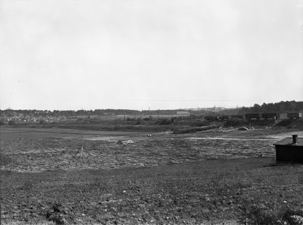 Vy österut över Årstafältet med järnvägsvagnar på Södra Stambanan.  
Till vänster Årsta Koloniträdgårdar framför Årstaskog där Högalidskyrkans torn sticker upp över träden.
Mitt i bilden en av ladugårdsbyggnaderna vid Ersta gård och till höger skogsområdet för blivande bostadsområdet Östberga.