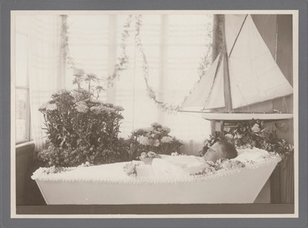 En död pojke ligger lite de parade i sin kista omgiven av blommor och en segelbåtsmodell .