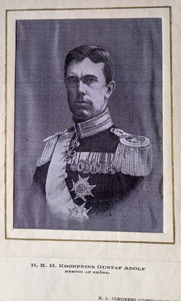 En vävd bild av kronprinsen Gustaf Adolf. Prinsen ser sträng ut. Han har sidbena. Han har på sig uniform med 7 medaljer på bröstet och en dekoration runt halsen. Han bär också två stora ordersstjärnor (troligtvis serafimerorden) och ett ordensband (serafimer). Prinsen har epåletter på axlarna med många fransar. 