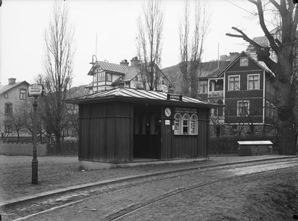 Liten träbyggnad med tre välvda fönster och en klocka på fasaden. I bakgrunden större trähus.