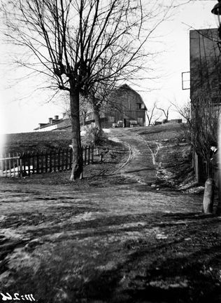 Högst upp på en kulle ligger ett gammalt trähus. Några träd i backen, till höger skymtas ytterligare ett hus.