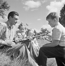 Kaknäs. Riksskyttet. En man sitter i gräset med ett gevär, omgiven av barn