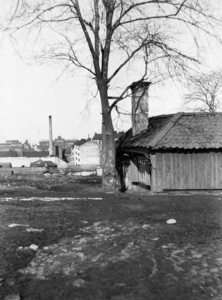 I förgrunden ett gammalt trähus med en hög skorsten. Bredvid står ett högt träd. I bakgrunden syns diverse bebyggelse och en hög fabriksskorsten.