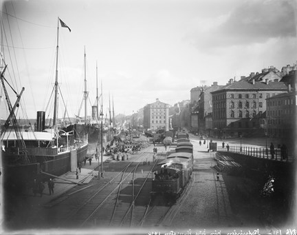 Svartvit bild med båtar, tåg och byggnader. I främre delen av bilden syns tågspår och flera vagnar. Till vänster ligger båtar i hamn.
